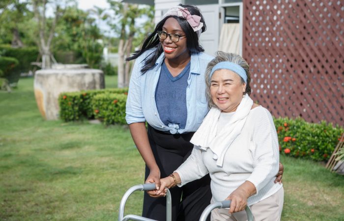 asian senior mother walk with walker and african american carer support in garden at home. old woman disabled walking and black caregiver young woman helping in back yard park outdoors. health care