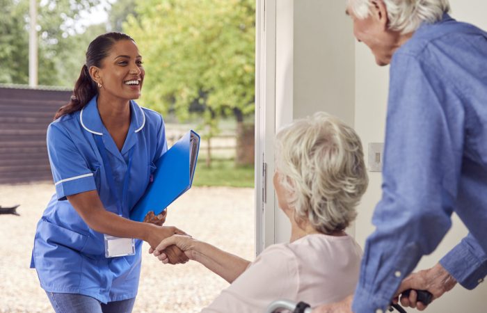 Senior Couple With Woman In Wheelchair Greeting Nurse Or Care Worker Making Home Visit At Door