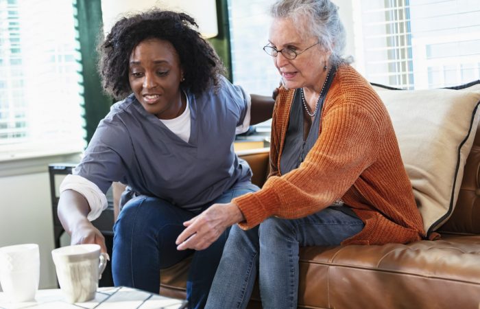 A senior woman in her 80s sitting at home on the living room sofa with a home healthcare aide who is helping her pick up a cup from the coffee table. The healthcare worker is an African-American woman in her 30s.