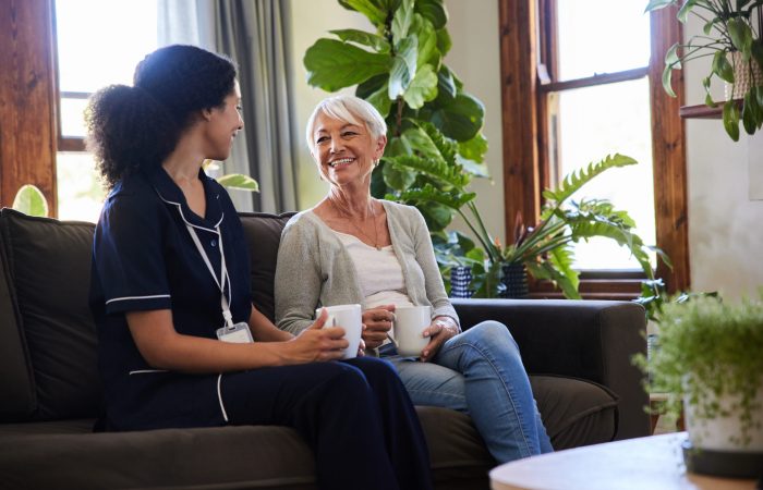 Smiling senior woman talking with a healthcare worker over coffee while sitting together on a living room sofa during a home visit